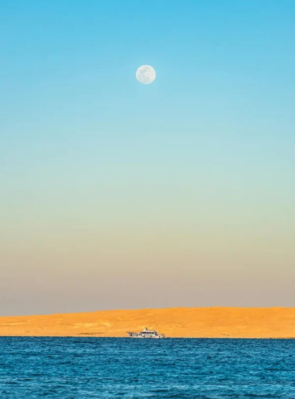 A serene view of the Red Sea in Hurghada, Egypt, with a full moon rising above golden sand dunes.