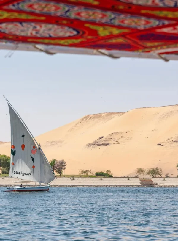 A traditional sailboat sails on the Nile with sand dunes in the background, under a vibrant canopy.
